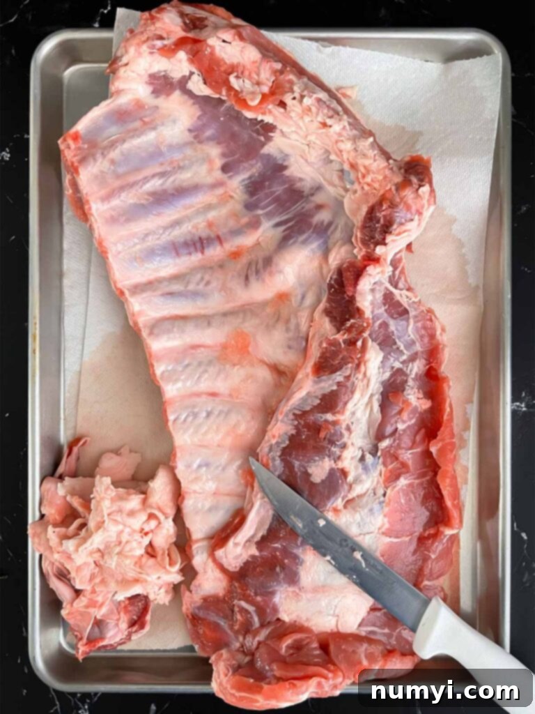A rack of spareribs on a baking sheet being trimmed of excess fat.
