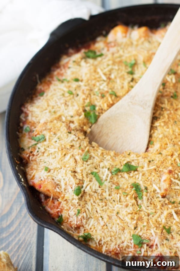 A large cast iron skillet full of chicken parmesan pasta with a golden breaded topping and a wooden spoon resting inside it