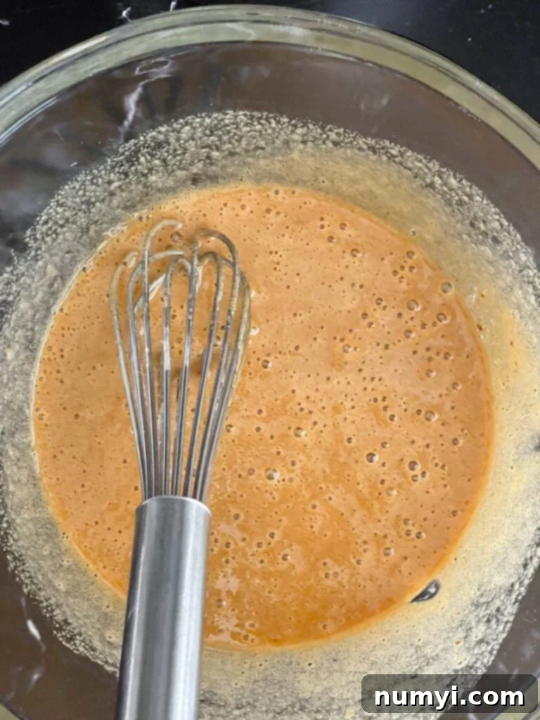 A large glass bowl holding a golden, slightly frothy mixture of wet ingredients, including whisked sugar and eggs, waiting for further additions like butter and buttermilk.