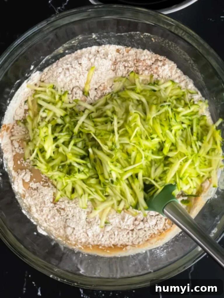 Freshly shredded zucchini being gently folded into the muffin batter with a spatula, demonstrating the incorporation of the key vegetable ingredient.