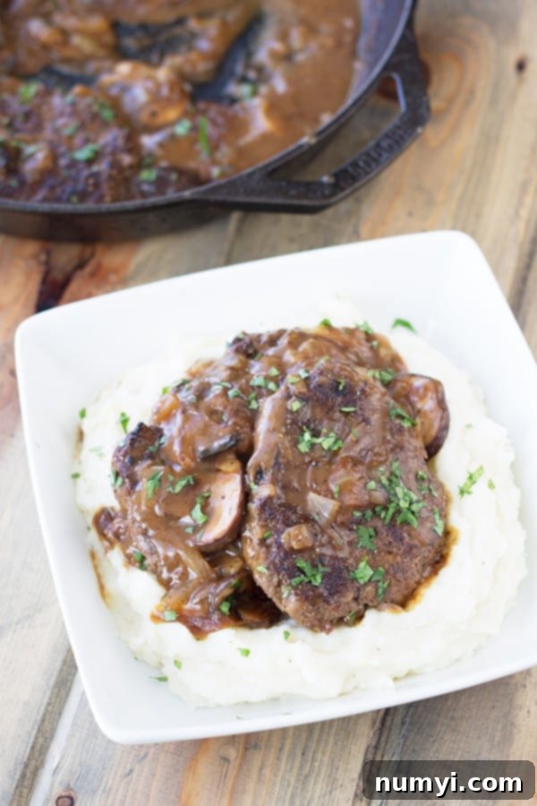 skillet full of salisbury with gravy in the background with a plate of mashed potatoes and salisbury steak with mushrooms in the foreground