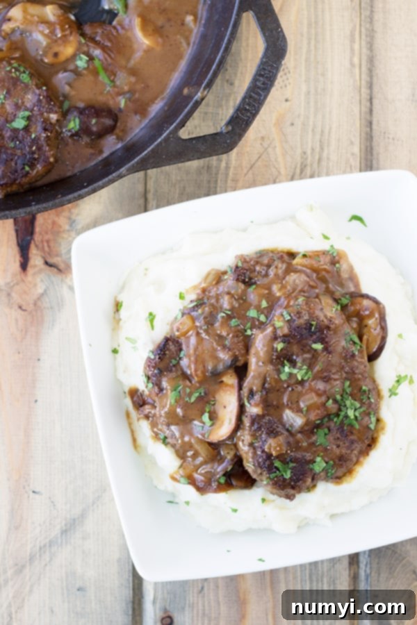 a photo shot from above with a corner of the skillet full of salisbury steak in the left corner, and a white dish of salisbury steak and mushrooms served over mashed potatoes in the middle right side