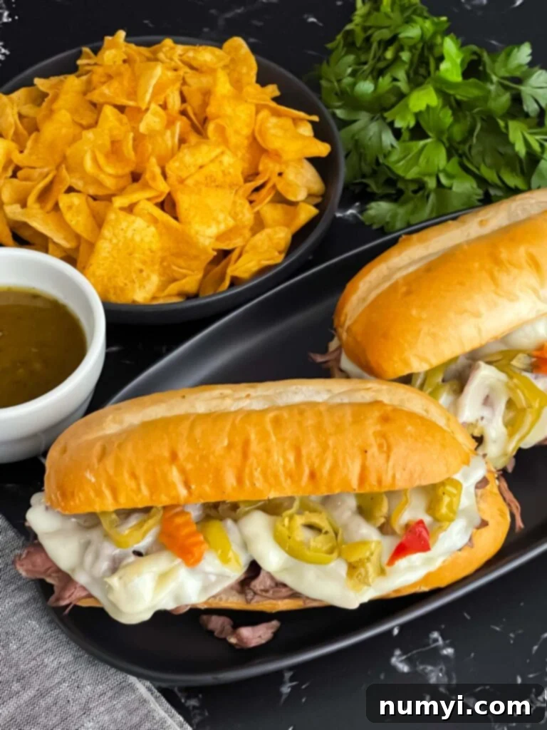 Melt-in-Your-Mouth Slow Cooker Italian Beef Sandwiches 2 Italian beef sandwiches on a dark plate, garnished with peppers and melted cheese, ready to be served.