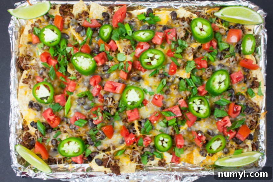Freshly baked Sheet Pan Nachos topped with diced tomatoes, green onions, sliced jalapenos, and lime wedges.
