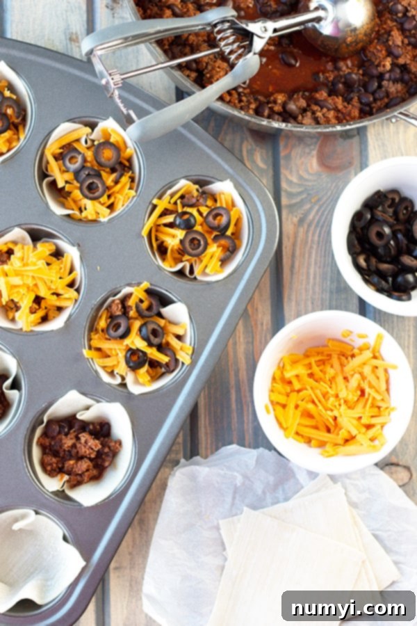 A bowl of beef enchilada mixture along with bowls of cheese and olives beside a cupcake tray housing unbaked enchilada cups ready to be cooked