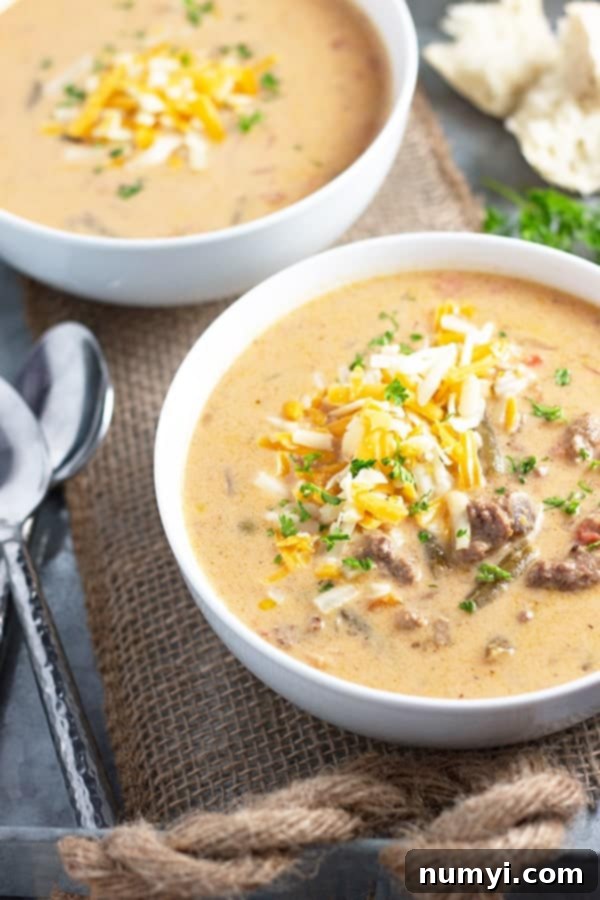 two white bowls of cheeseburger soup with cheese and garnish on top sitting on a jute-type fabric beside two spoons