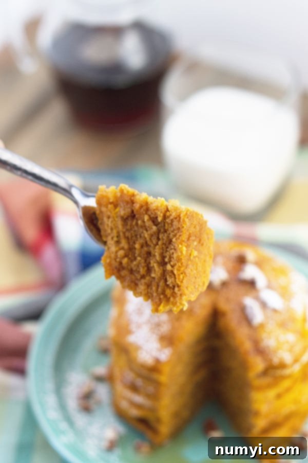 Ultimate Pumpkin Spice Pancakes 4 A stack of golden pumpkin pancakes in the background, slightly out of focus, with a single bite skewered on a fork in sharp focus in the foreground, ready to be enjoyed.