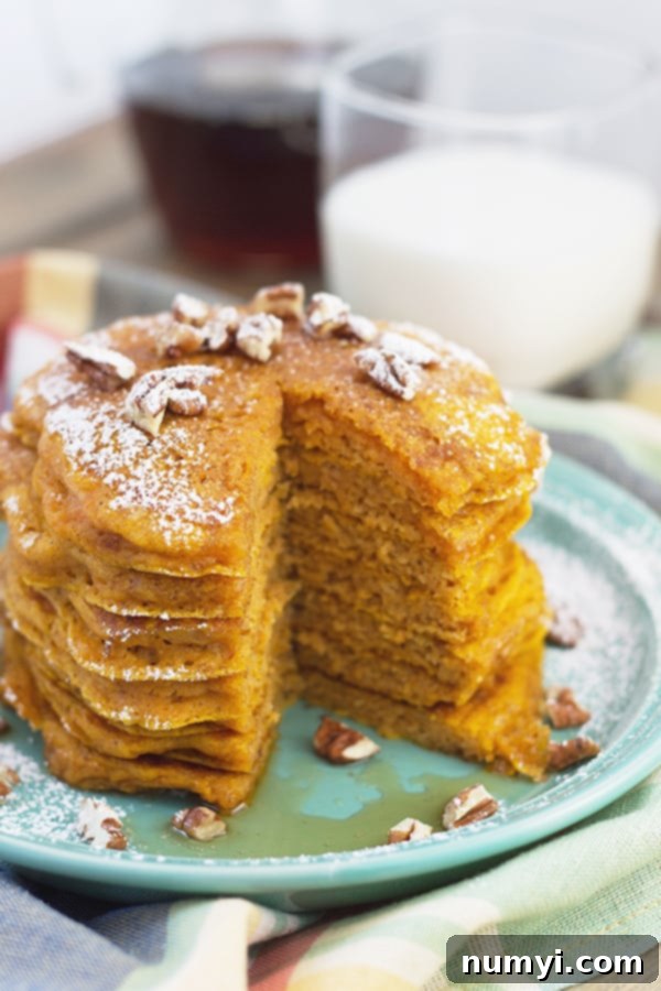 Ultimate Pumpkin Spice Pancakes 5 A stack of fluffy pumpkin pancakes, artfully arranged and topped with powdered sugar, chopped nuts, and a glistening stream of maple syrup. A slice has been taken out, revealing the moist interior.