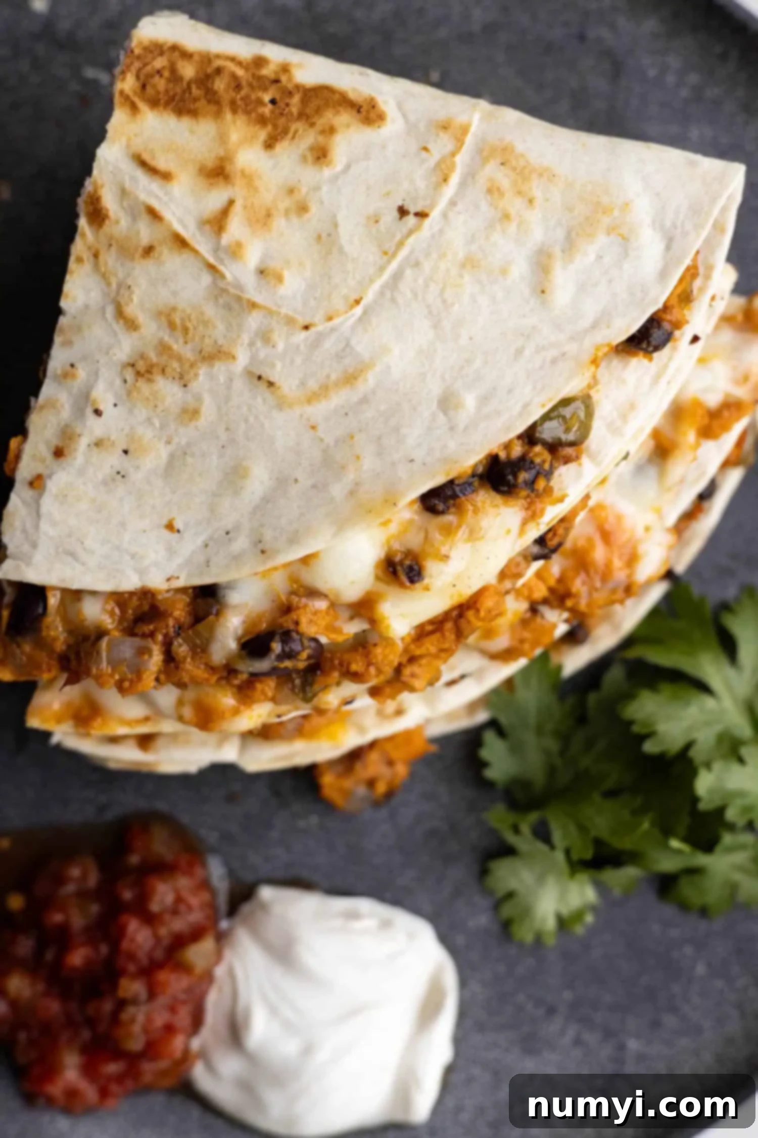 Overhead view of a stack of golden brown black bean sweet potato quesadillas, garnished with fresh cilantro, with small bowls of sour cream and vibrant salsa on the side.