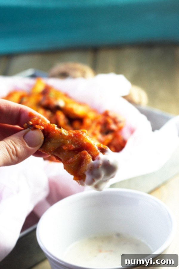 A single baked sriracha chicken wing, perfectly crispy, dipped in a creamy ranch dressing, with a basket of more wings blurred in the background, highlighting the delicious serving suggestion.