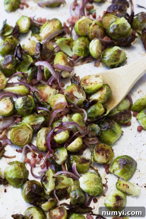 Oven-Roasted Brussels Sprouts with Ham & Caramelized Onions 3 A wooden spoon stirring a baking sheet filled with perfectly roasted Brussels sprouts, savory ham pieces, and softened, caramelized red onions.