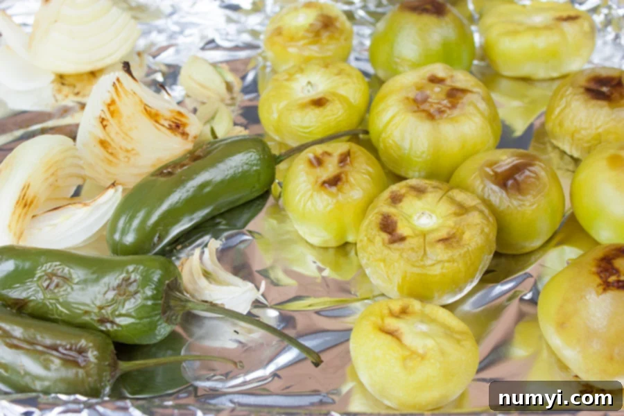 Roasted tomatillos, jalapeños, and onions on a foil-lined baking sheet after baking, showing their softened and slightly charred appearance.