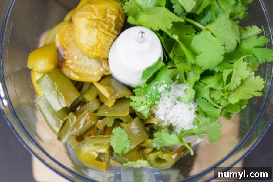 Roasted tomatillos, jalapeños, and onions are placed into the bowl of a food processor, ready to be blended into salsa verde.