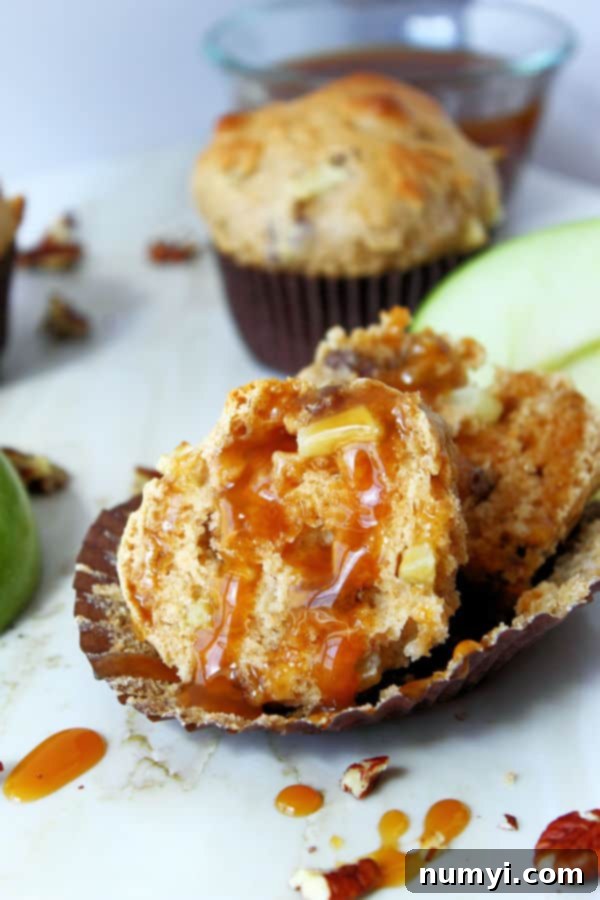 An apple muffin baked with pecans, cut open in its liner, revealing the soft interior and apple chunks, beautifully drizzled with glistening caramel sauce.