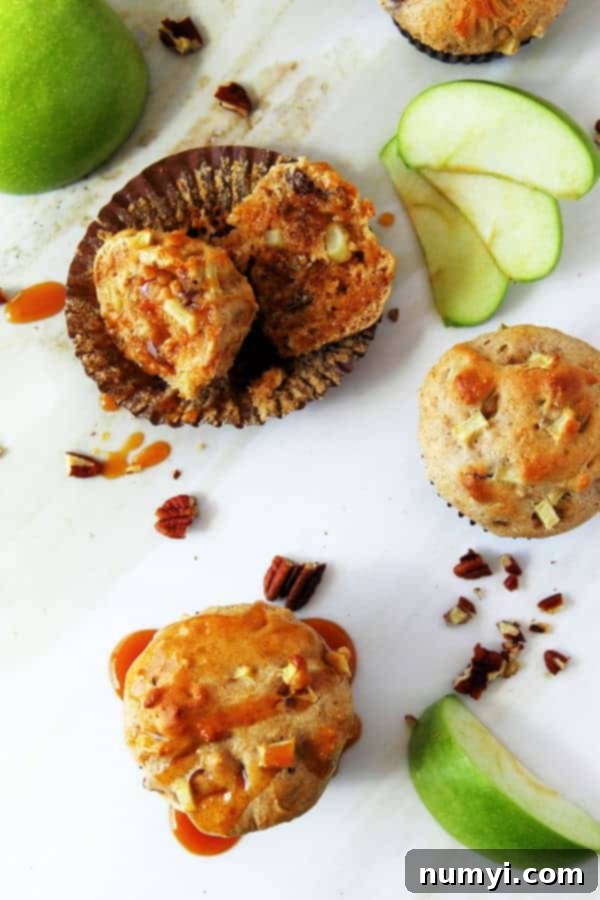 A cluster of apple muffins with pecans on a white surface. One muffin is cut open in its paper liner and generously drizzled with caramel sauce, surrounded by fresh Granny Smith apple slices and scattered chopped pecans.