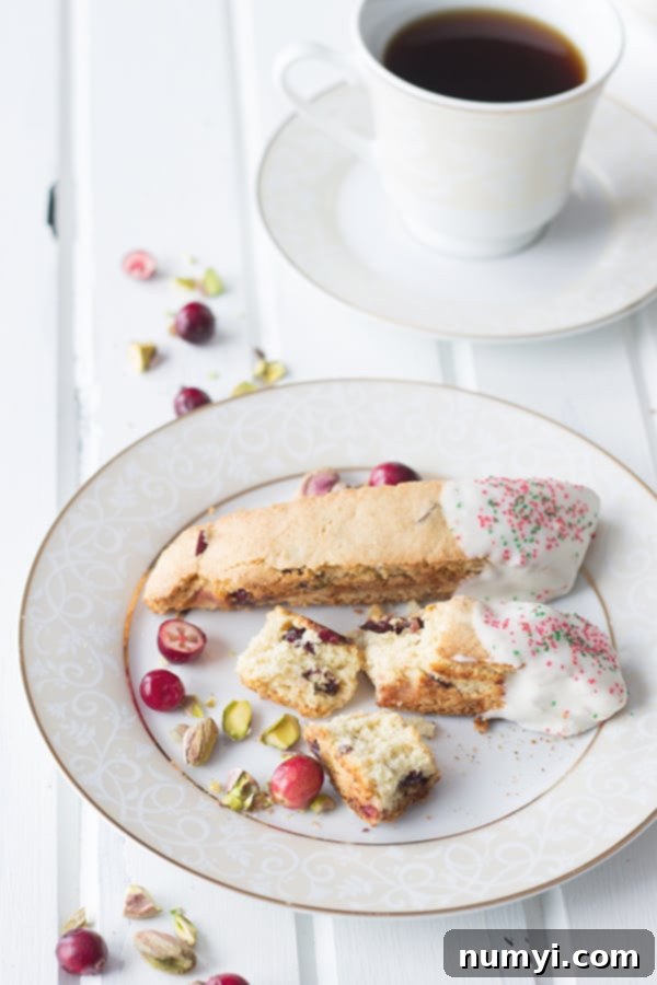 Crunchy Cranberry Pistachio Delight 3 A white plate showcasing two exquisite cranberry pistachio biscotti, one whole and one broken to reveal its tempting, crunchy interior filled with red cranberries and green pistachios. Some crumbs are scattered around.