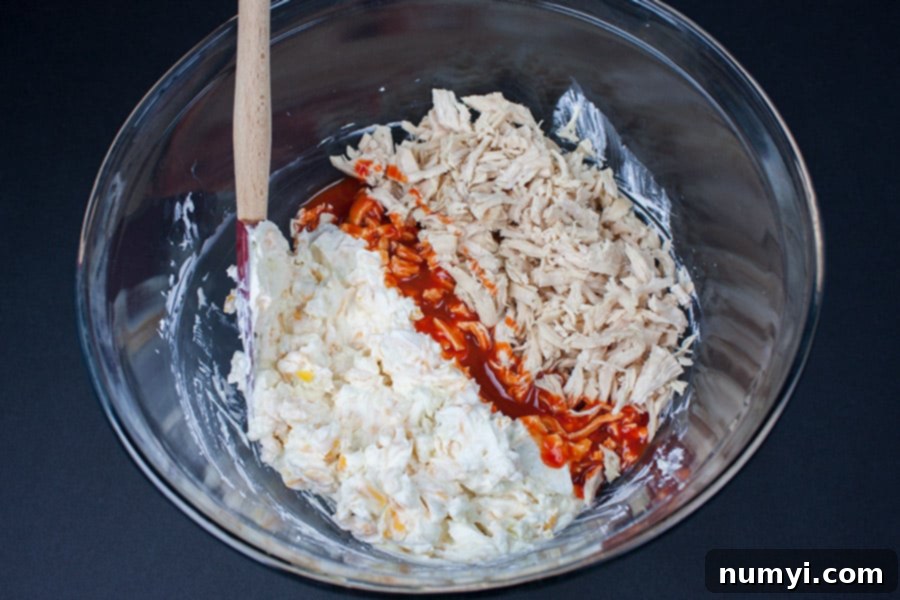 Shredded chicken, cream cheese, blue cheese, cheddar cheese, and sour cream in a glass mixing bowl.