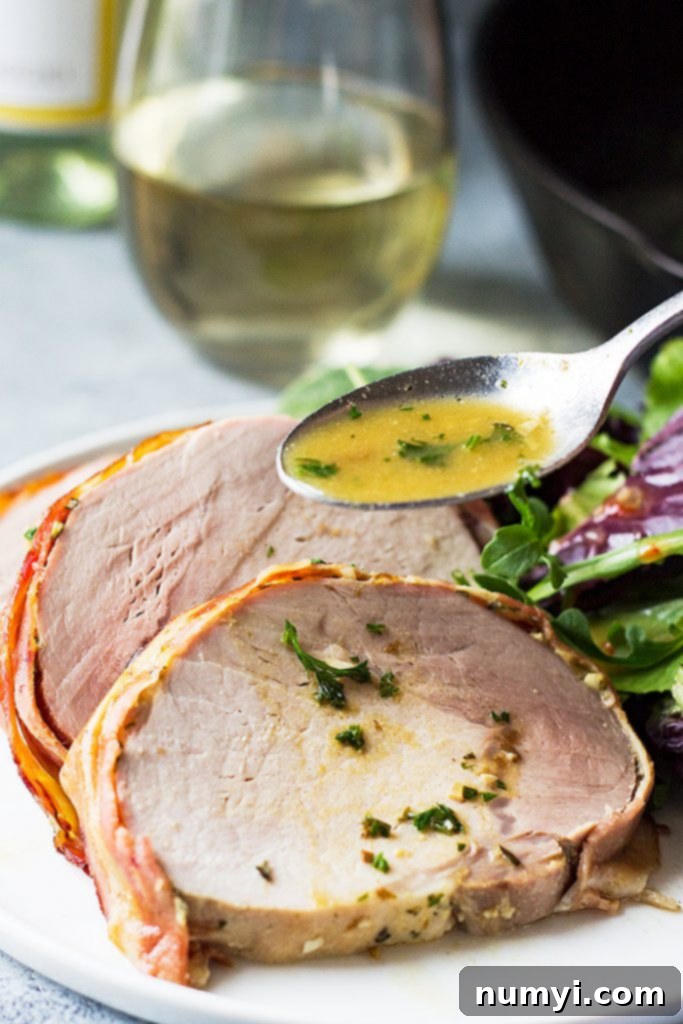Crisp Bacon-Wrapped Pork Loin 5 A hand spooning rich white wine sauce over neatly sliced portions of bacon wrapped pork loin on a plate.