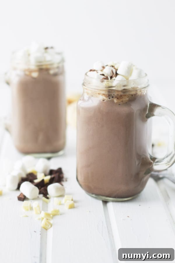 two mason jar mugs full of creamy hot chocolate with marshmallows on top and around on the table