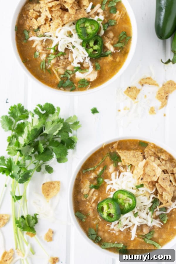 Slow Cooker White Chicken Chili 3 Two bowls of Slow Cooker White Chicken Chili on a white table, richly garnished with avocado slices, sour cream, shredded cheddar cheese, fresh cilantro, and a side of tortilla chips and fresh jalapeño slices.