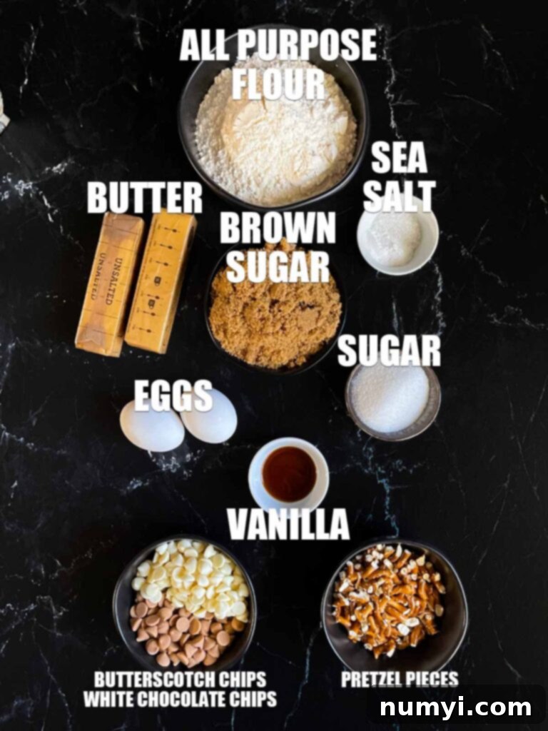Ingredients for salted butterscotch pretzel cookies laid out on a dark surface, including flour, sugars, butter, eggs, vanilla, pretzels, and chocolate chips.