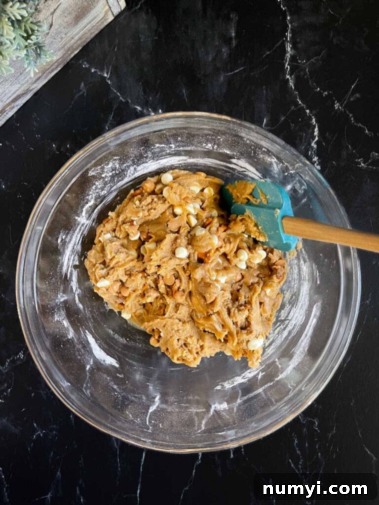 Salted Butterscotch Pretzel Cookie dough, with chips and pretzels folded in, resting in a glass bowl on a dark surface.
