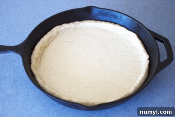 A cast iron skillet prepared with pizza dough, showing the crimped crust edge and fork marks on the bottom, ready for toppings.