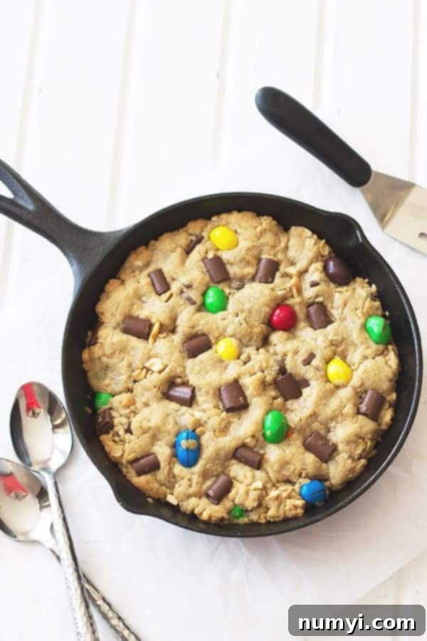 An overhead shot of a warm, gooey Monster Skillet Cookie in a cast iron pan, laden with colorful M&Ms, melty chocolate chunks, and roasted peanuts, ready to be served.