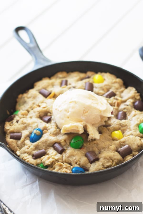 A warm Monster Skillet Cookie in a cast iron skillet, topped generously with a melting scoop of vanilla ice cream, creating a mouthwatering dessert experience.