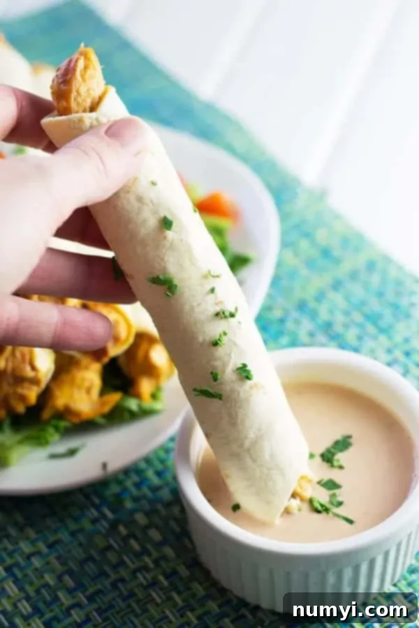 Close-up of a hand dipping a crispy baked chicken taquito into a small ramekin of creamy dipping sauce.