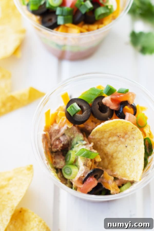 an overhead photo of a couple of plastic cups full of seven layer taco dip with chips in one and more scattered around