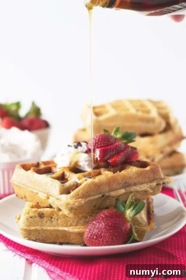pouring syrup over a stack of 2 strawberry chocolate chip waffles with whipped cream and berries on top, a truly indulgent breakfast