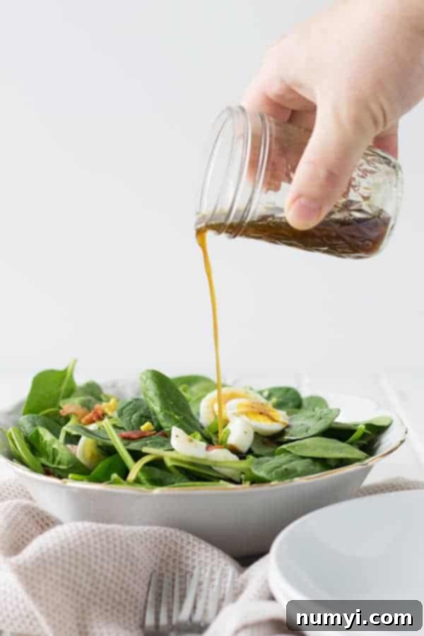 Side view of warm spinach salad with bacon with bacon dressing being poured from a mason jar above the salad.