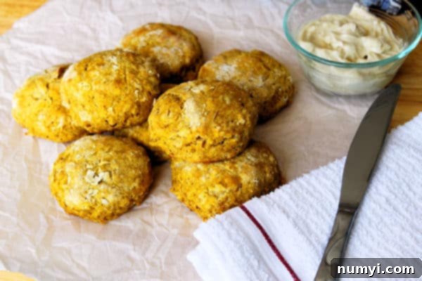 Pumpkin Biscuits stacked on top of each other on a piece of parchment with maple butter, a knife, and a dish towel to the right