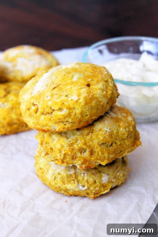 three pumpkin biscuits stacked on top of each other in the foreground with more biscuits and a glass bowl full of maple butter in the background