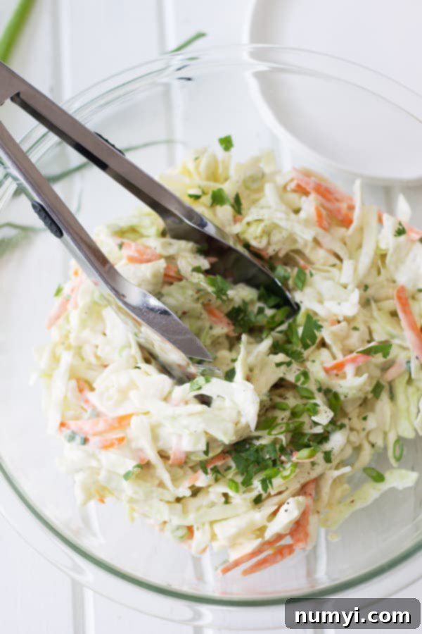 Shredded cabbage and carrot coleslaw in a clear glass mixing bowl with serving tongs, ready to be transferred to plates.