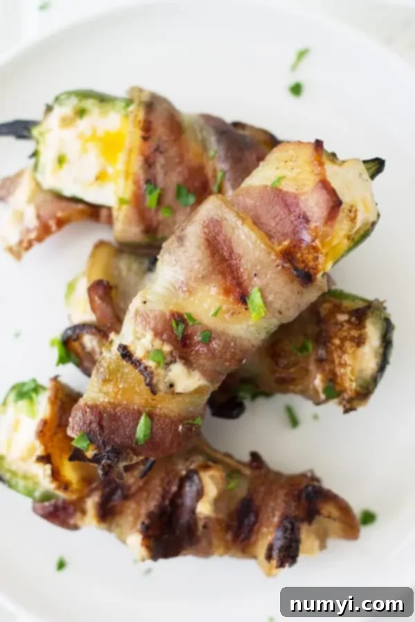Closeup overhead shot of grilled jalapeno poppers, filled with creamy cheese and wrapped in bacon, garnished with fresh herbs on a plate.