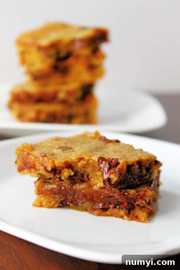 Two perfectly baked Banana Chocolate Chip Peanut Butter Blondies, golden brown and studded with chocolate, stacked on a white plate in the foreground, with more delicious blondies blurred in the background.