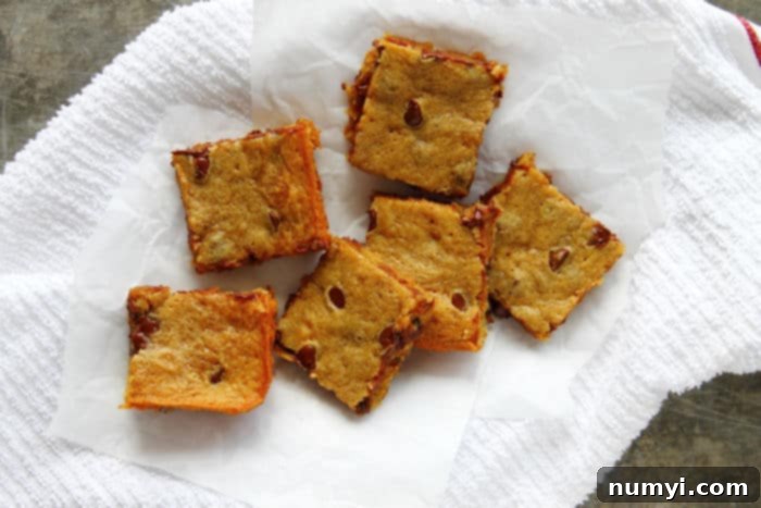 Six square-cut Banana Peanut Butter Chocolate Chip Blondies, neatly arranged on a rustic white tea towel, showcasing their golden brown edges and melty chocolate chips.