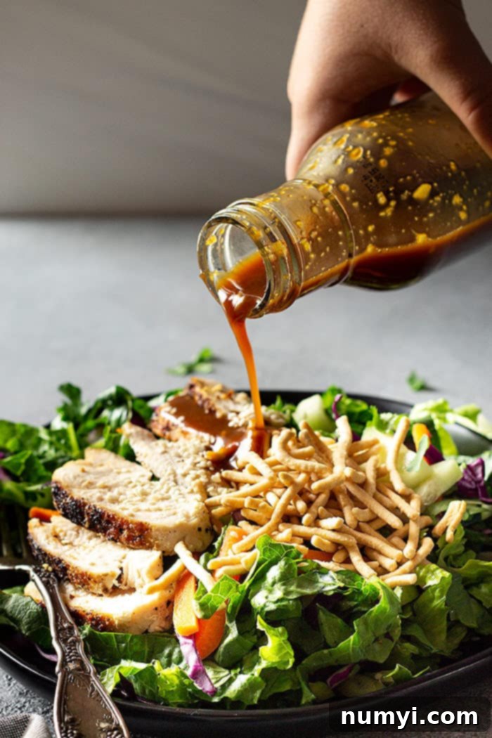 A hand drizzling the delicious homemade sesame ginger Asian dressing over a freshly assembled Asian Chicken Salad, showing the rich, savory sauce cascading over the vegetables and chicken.