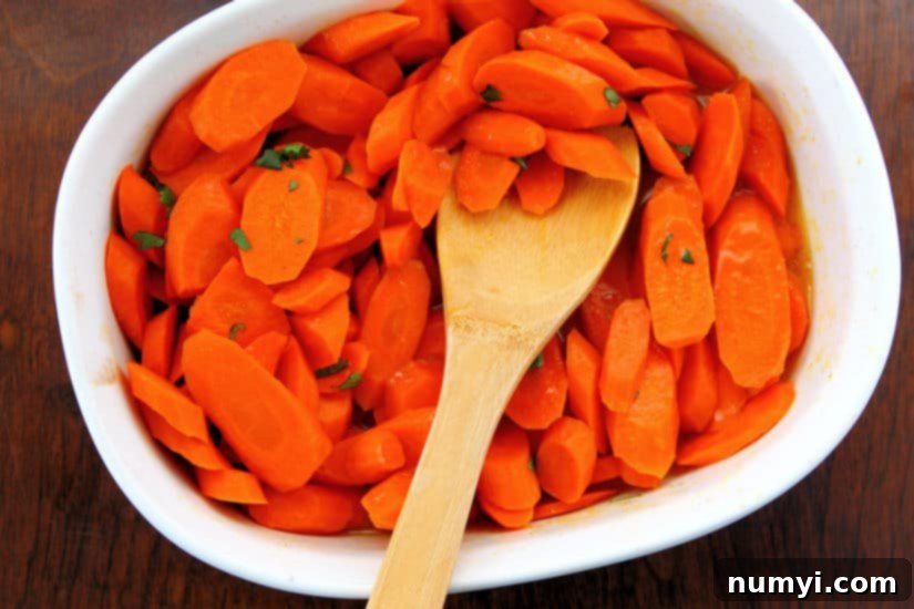 Overhead shot of gleaming ginger maple glazed carrots in a white serving bowl, garnished lightly with fresh herbs, alongside a wooden spoon.