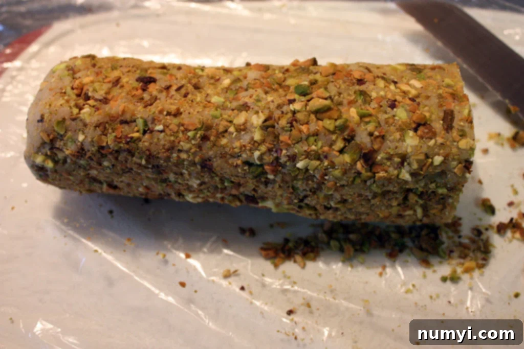 Cookie dough log evenly coated with finely chopped pistachios, resting on plastic wrap.