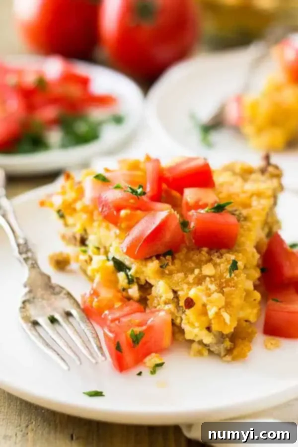 A close-up shot of a single slice of hash brown pie on a plate, garnished with fresh chopped tomatoes, revealing the layers of sausage, cheese, and potatoes, with a fork ready to dig in.