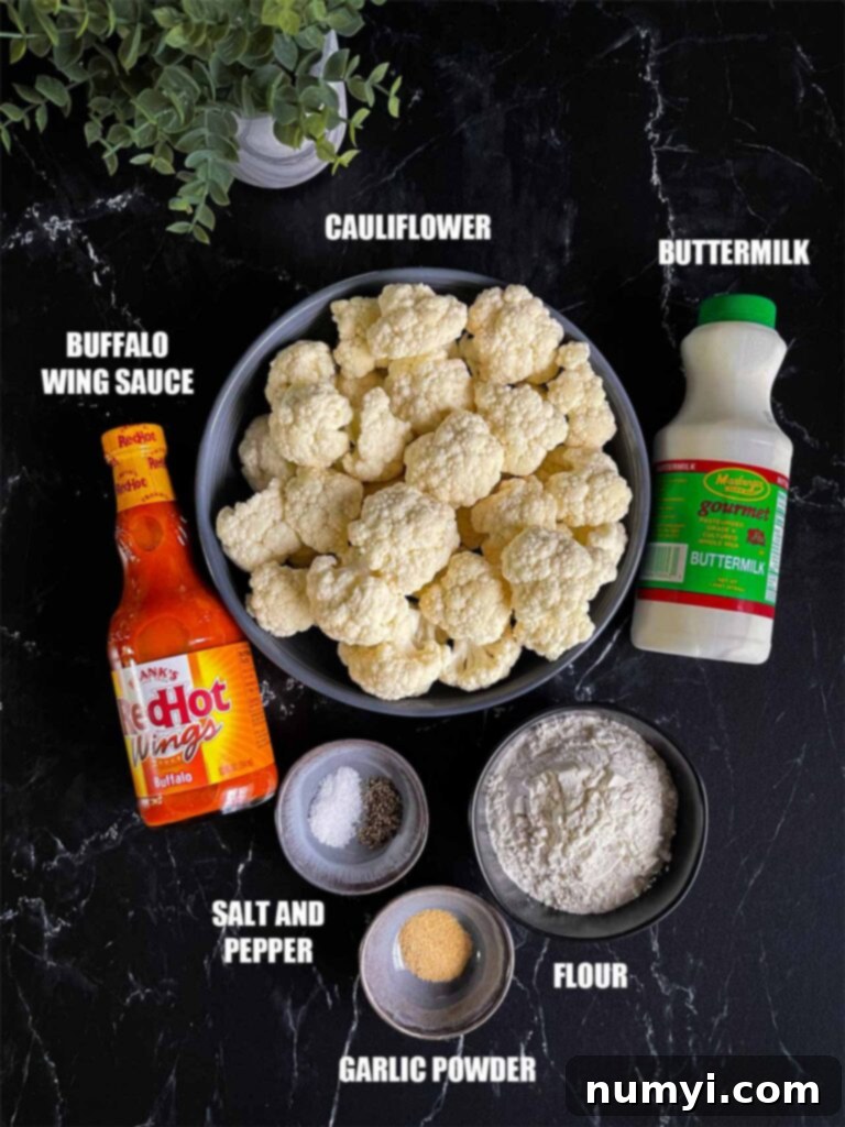 A perfectly arranged flat lay of fresh ingredients required for making baked buffalo cauliflower bites, including a whole head of cauliflower, all-purpose flour, buttermilk, garlic powder, salt, pepper, and a bottle of buffalo sauce, laid out on a dark, rustic surface.