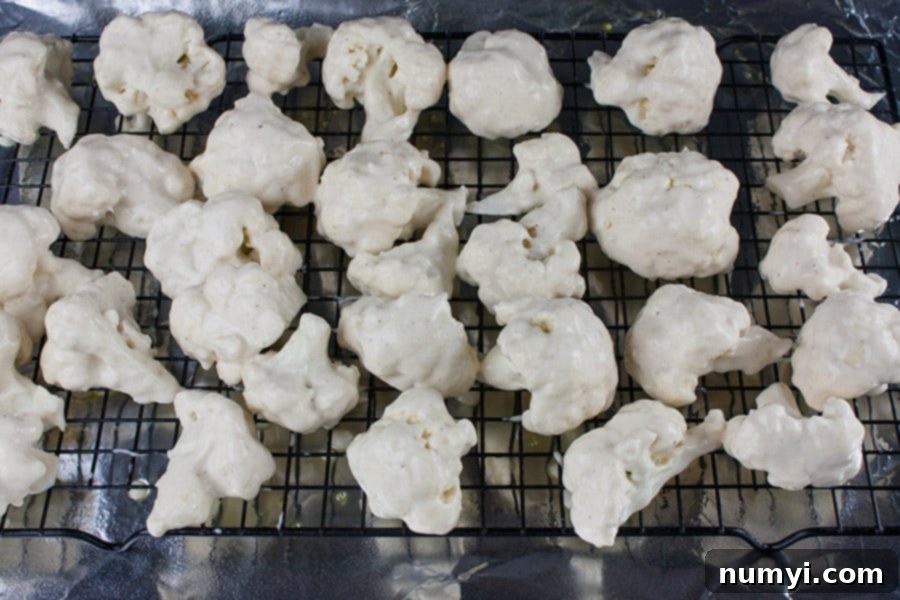 Raw cauliflower florets, uniformly coated in a smooth, light-colored batter, neatly arranged in a single layer on a wire rack which rests on a baking sheet, prepped and ready for baking.