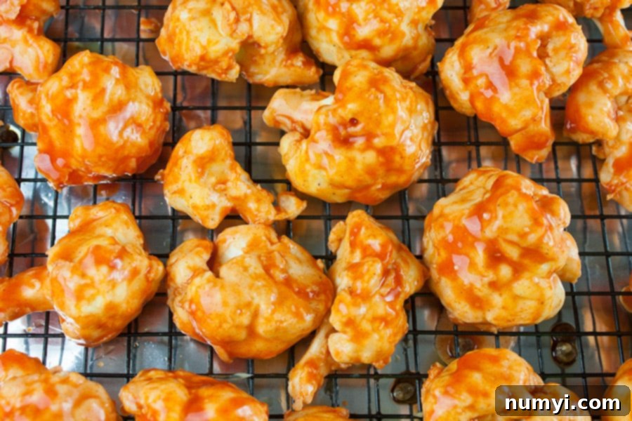 Freshly baked cauliflower bites, now glistening with a rich coating of vibrant buffalo sauce, neatly arranged on a wire rack set within a baking sheet, poised for their final bake to achieve ultimate crispiness and flavor.