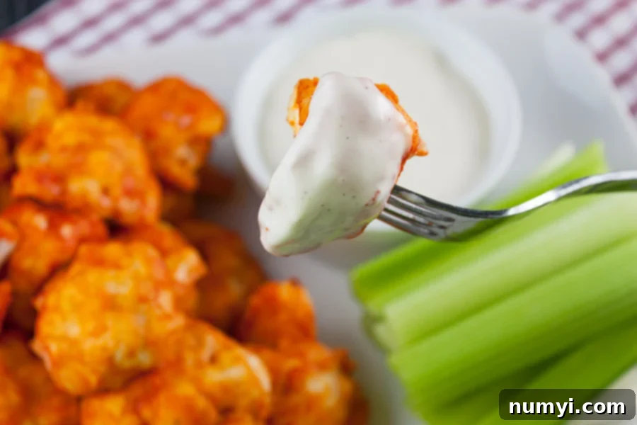 A single, perfectly baked buffalo cauliflower bite is delicately held on a fork, lavishly dipped into a pool of creamy ranch dressing, highlighting its appealing texture and generous coating of sauce.