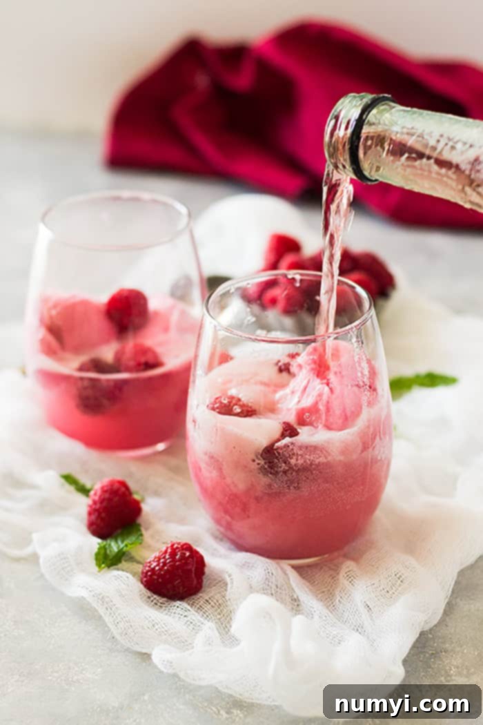 Two glasses filled with raspberry sherbet and fresh raspberries, with pink champagne being poured into them, creating a bubbly dessert drink.
