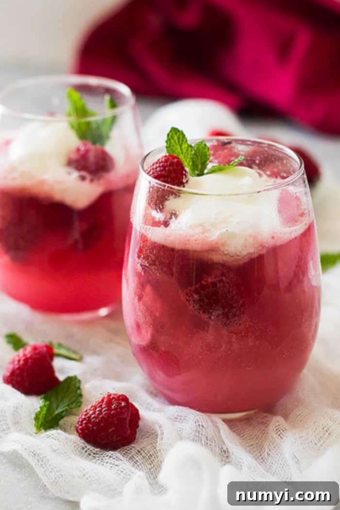 Two full glasses of sparkling pink champagne floats, topped with creamy whipped cream and fresh raspberries, ready to be enjoyed.