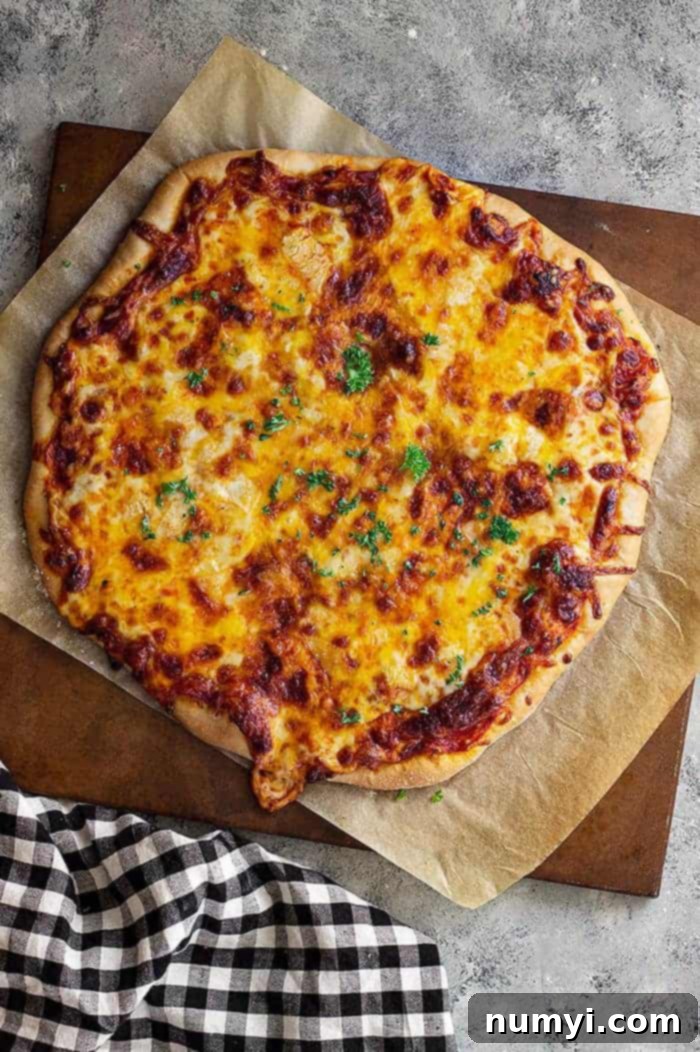Perfectly Homemade Cheese Pizza 7 overhead: homemade cheese pizza on parchment paper on a wooden cutting board with a black and white checkered towel to the left